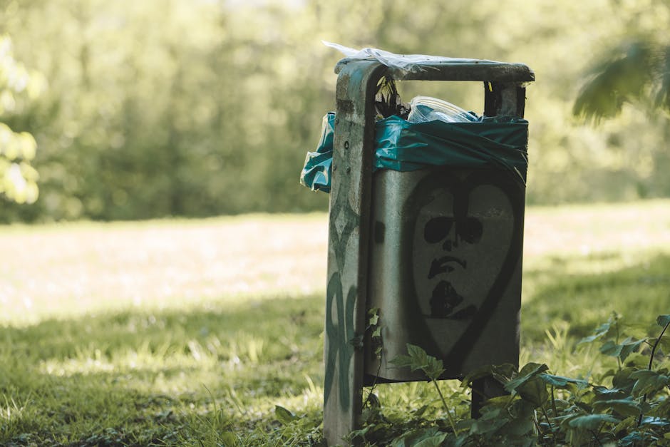 Two large black plastic garbage bags filled with waste are positioned on the edge of a sidewalk next to a black metal fence. The bags appear to be tightly packed and somewhat crumpled, with visible cr
