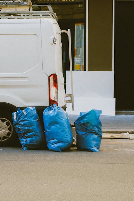 The image shows the rear of a white commercial van parked on a paved street in front of a building with large glass windows and a white panel. Three blue rubbish bags, made of plastic with a slightly 