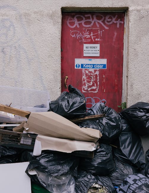 The image shows the rear of a white commercial van parked on a paved street in front of a building with large glass windows and a white panel. Three blue rubbish bags, made of plastic with a slightly 